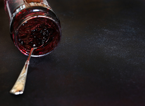 Spoon with berry jam resting on an almost empty glass jar isolated on dark background. Overhead view, copy space.