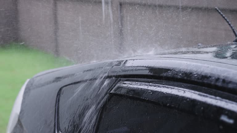 SLO MO: torrential rain falling heavily on a beautiful black car parking in front of a house in a garage but exposing its trunk outdoors.