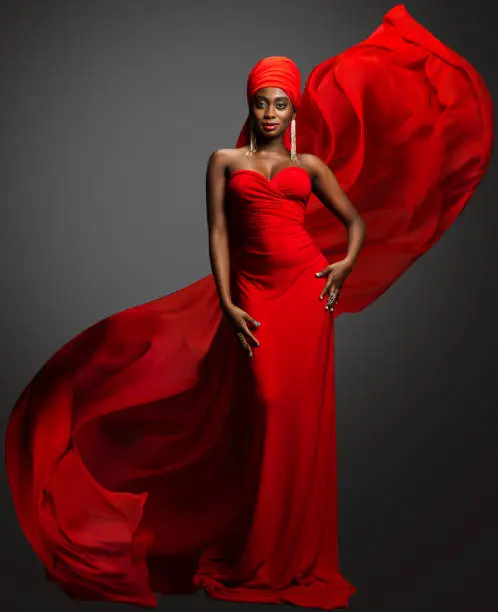 Photo of Fashion Woman in Red Dress and Hijab. African Model in Evening long Gown with flowing Silk Fabric over Dark Gray background. Muslim Bride in Wedding chiffon Veil