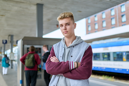 Portrait of young confident guy at railway station of electric city commuter train. Teenage male student 18, 19 years old looking at the camera with crossed arms. Youth, urban lifestyle