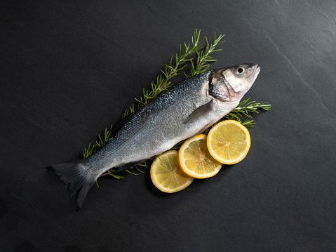 Raw sea bass on dark stone background with lemons and rosemary.