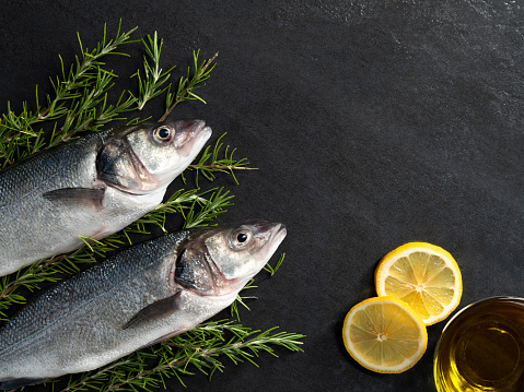 Two raw sea basses on black stone background with rosemary, lemon and olive oil.