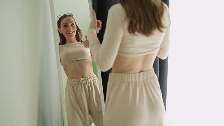 A young woman wearing oversized pants after losing weight looks at her figure in front of the mirror, very pleased that she managed to lose weight.