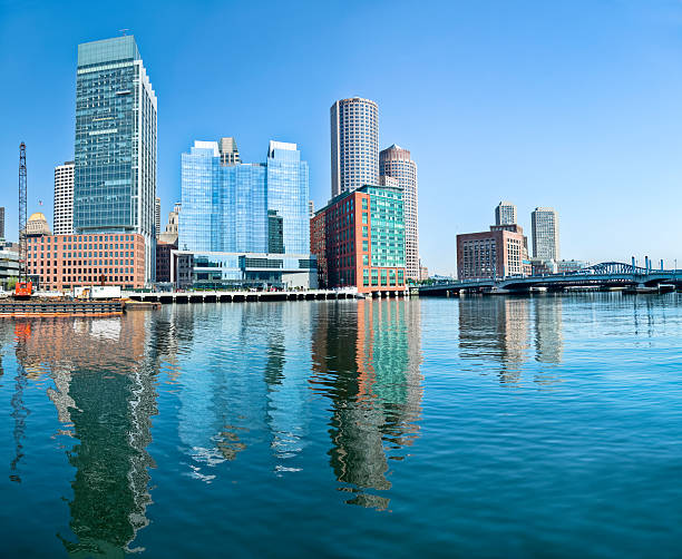 The Boston skyline along the Fort Point Channel contains numerous skyscrapers and buildings of various shapes, heights, and architecture. They each cast a reflection on the rippling waters of the channel under a clear blue sky.