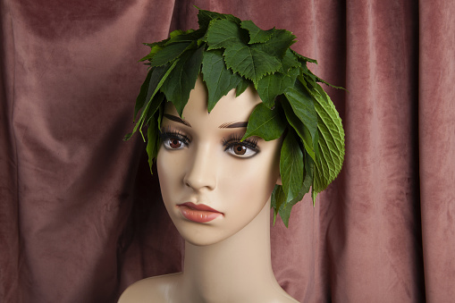 a display mannequin wearing hair of leaves and representing a nature goddess in front of a pink velvet curtain. Minimal color still life photography.