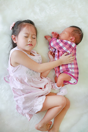 Asian older sister and newborn baby boy sleeping together on white fur background.
