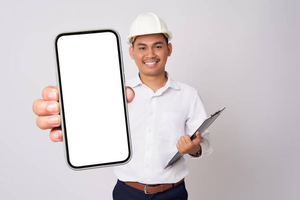 istock Smiling young Asian architect engineer employee man in a formal shirt helmet holding smartphone with blank screen and clipboard isolated on white background. People work on architecture project 1677670849