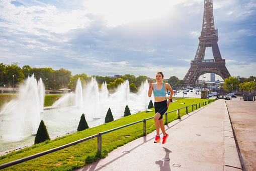 Athletic young woman in sports outfit mid-air, running down the pedestrian lane by the fountains near the Eiffel tower in Paris on a sunny summer day