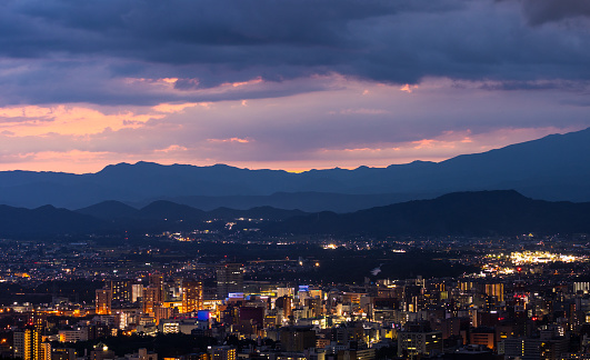 A high-angle view over Morioka City at night at sunset with the distant mountains of Shizukuishi visible in the background.