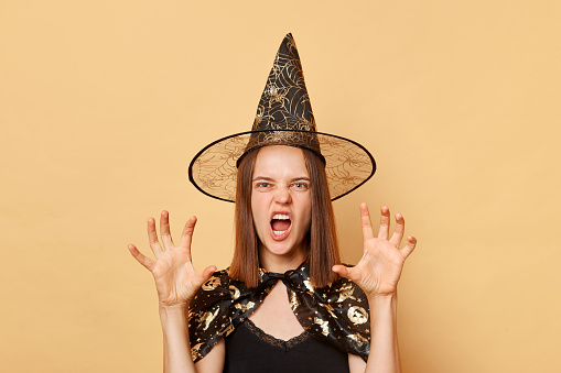 Scary young woman sorcerer wearing witch costume and carnival cone hat celebrating halloween isolated over beige background raised her arms paws grimacing