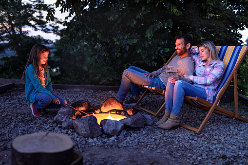Happy parents and their small girl enjoying while spending a night by the campfire in the backyard.