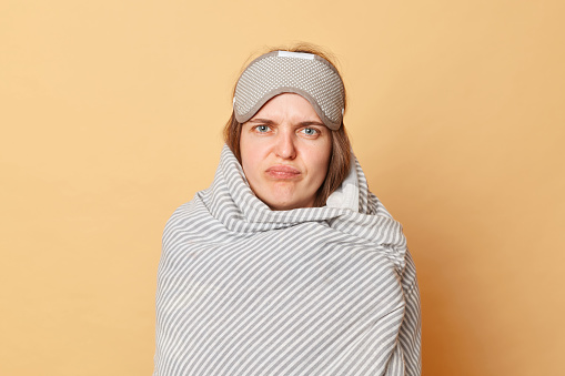 Sad unhappy upset woman wrapped blanket and wearing sleeping eye mask isolated over beige background standing with frowning face grimacing do not want to wake up