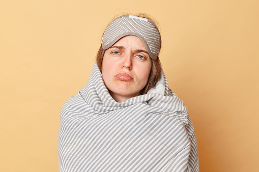 Unhappy sad woman wrapped blanket and wearing sleeping eye mask isolated over beige background looking at cmaera with pout lips lazy to wake up early.