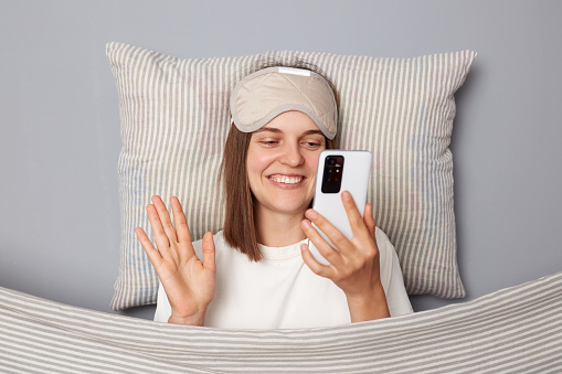 Cute smiling woman in white T-shirt and sleeping eye mask lie in bed on pillow under blanket isolated on gray background having video call waving hand hello wakeup blog.