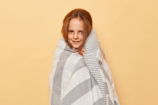 Cute charming smiling little girl with blanket standing isolated over beige background posing wrapped in plaid wake up early morning.
