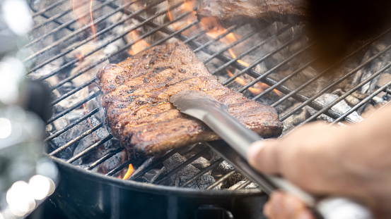 Close-up of person's hand picking up roasted meat with pair of tongs on hot barbecue grill.