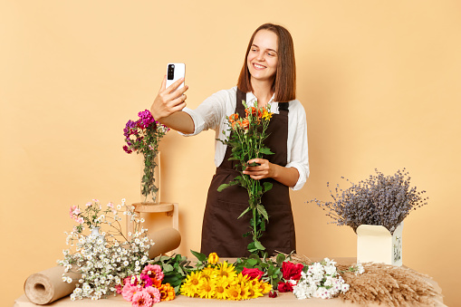Beautiful woman florist blogger making selfies for followers retail seller offering buyer big choice of flowers in shop room wearing brown apron standing against beige wall.