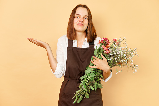 Uncertain woman florist wearing brown apron holding bouquet of flowers standing isolated over beige background shrugging shoulders looking at camera with confused expression.
