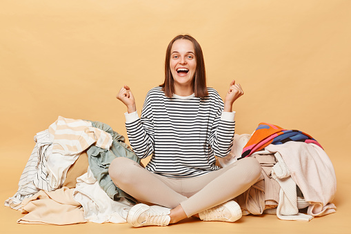 Overjoyed Caucasian woman posing near heap of multicolored unsorted clothes isolated over beige background clenched fists rejoicing buying lots new attires.