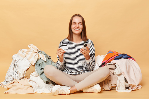 Smiling Caucasian woman posing near heap of multicolored unsorted clothes isolated over beige background buying attires online holding smartphone and credit card entering data.