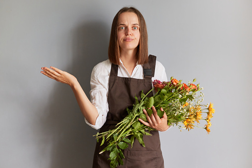 Puzzled confused female florist wearing brown apron holding flowers isolated over gray background spreading hand shrugging shoulders looking at camera with uncertain face.
