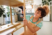 Young beautiful casually clothed woman assembling shelf with electric drill at home