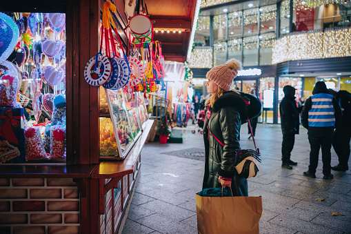 Woman in jacket and bobble hat standing in front of Christmas street market stand with candy and toys, looking at the offer