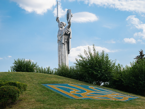 Kyiv, Ukraine -August 27, 2023: Ukrainian Motherland Monument against a sky with clouds and the state emblem of Ukraine in the foreground