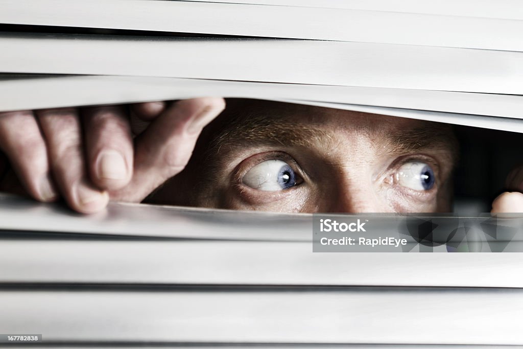 Fearful man looking sideways through venetian blind This terrified man looking out to the side  through a venetian blind could be witnessing a crime, watching the suspicious behavior of neighbors, or simply a paranoid, isolated shut-in.   Paranoia Stock Photo