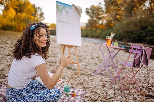 Young artist sitting on blanket near river and painting a image
