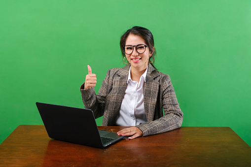 Successful happy Asian business woman sit work at office showing thumb up gesture isolated on green background