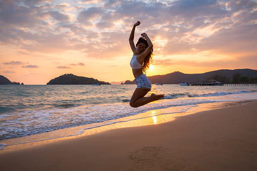 Summer woman vacations concept, Travel asia woman with white bikini and shorts jeans jumping on sea beach in sunset at Koh Mak, Thailand