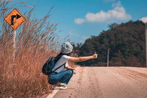 Back view of traveler asian woman with travel bag hitchhiking car on country road in Thailand