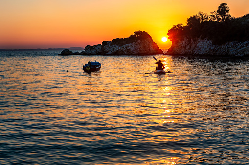 Silhouette man in a canoe rowing towards a beautiful sunset over the sea. The sun is setting between two rocks in the horizon, with a small motor boat anchored near by