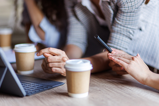Close up shot of small group of unrecognizable young businesswomen enjoying takeaway coffee while using smart phones, laptop and reviewing data on documents during a project brief.