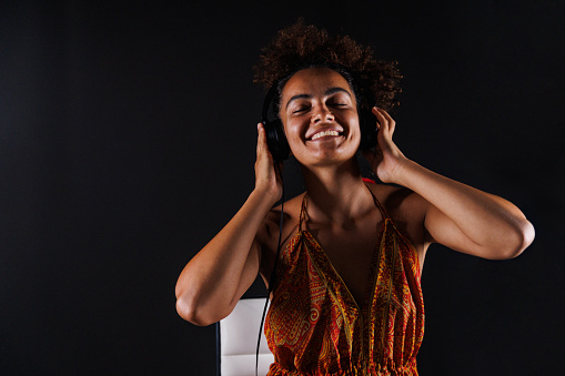 Portrait of happy young woman, with brown curly hair, sitting against black background, moving with the rhythm of her favorite song that she is listening to via headphones.