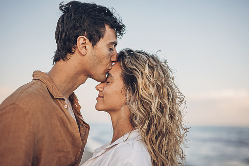 Man and woman, happy young couple standing on the beach by the sea. Man is kissing a woman.
