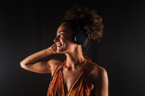 Portrait of cool young woman standing against black background and enjoying listening to music via headphones.