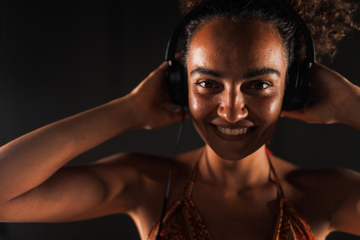 Headshot of cool young woman standing against black background and enjoying listening to music via headphones. She is looking at camera and smiling joyfully.