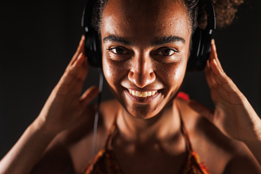 Headshot of cool young woman standing against black background and enjoying listening to music via headphones. She is looking at camera and smiling joyfully.