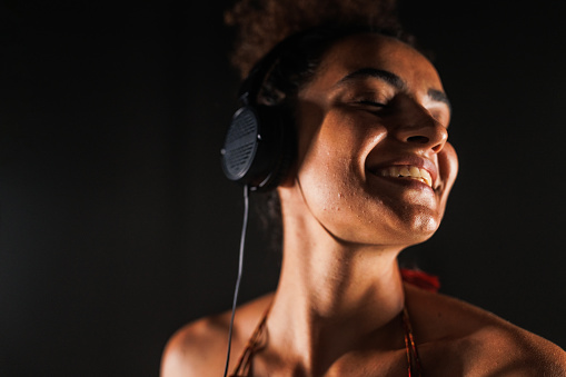 Headshot of cool young woman standing against black background and enjoying listening to music via headphones.