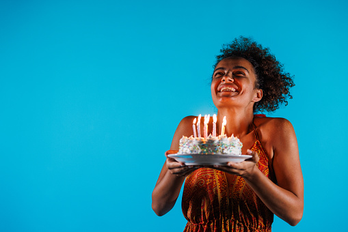 Portrait of cheerful young woman standing against blue background and holding a birthday cake with colorful candles and making a wish before blowing them out.