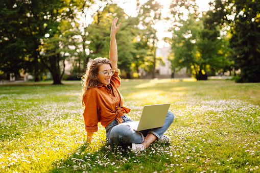 Young woman is sitting in a sunny park on a clearing and working with a laptop, blogging, online learning. Concept of remote work, freelancing, technology.