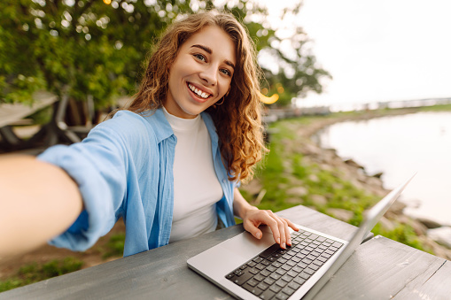 Happy woman in stylish clothes sitting at a table outdoors taking a selfie, working on a laptop with a beautiful view of the lake. Close up selfie of smiling female freelancer with laptop.