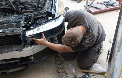 The car mechanic sits on his knees near the hood of a car. He is installing a new car headlight in his workshop.