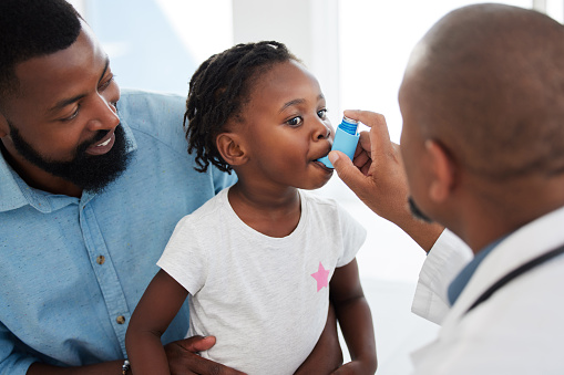 Doctor helping a child patient with an inhaler for asthma in his office at a medical clinic. Healthcare worker consulting a girl with chest or respiratory problems with pump in a children's hospital