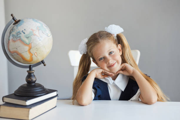 retrato de una hermosa niña con uniforme de moda y dos colas de caballo peinado se sienta en un escritorio en la lección en la escuela. linda y bonita colegiala caucásica sonríe y mira a la cámara. espacio de copia - portrait little girls 8 9 months hair bow fotografías e imágenes de stock