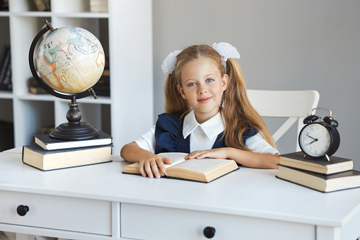 A beautiful schoolgirl in a uniform with two ponytails hairstyle is reading a book in the school library. Portrait of a cute girl in class with modern interior.