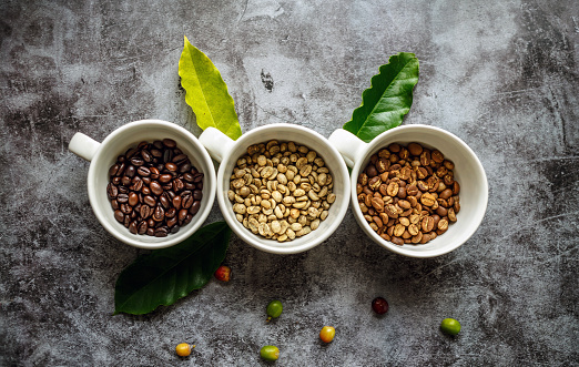Top view of many varieties of coffee beans available, including both fresh roasted and unroasted options in coffee cups, green coffee beans, and medium and dark roasted.