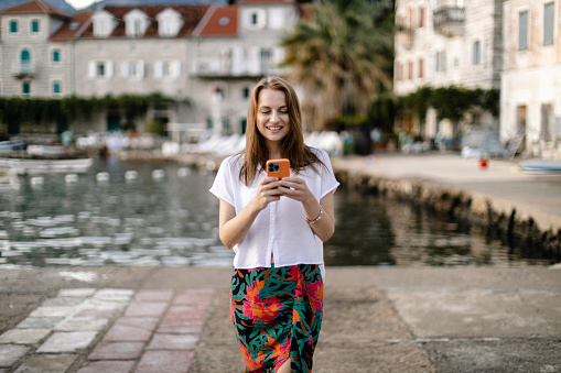 Smiling young woman using a smartphone and exploring an old Mediterranean town at the seaside on a sunny summer day